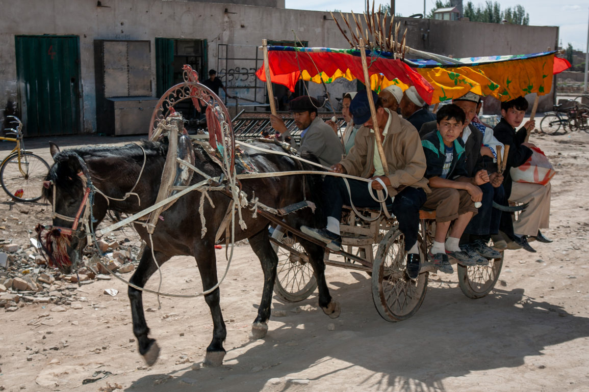 horse drawn cart on dusty unpaved street with at least 9 people aboard