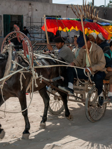 horse drawn cart on dusty unpaved street with at least 9 people aboard