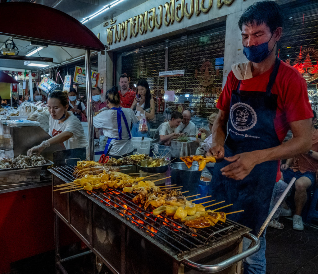 man grilling chicken over charcoal with another vendor in the background