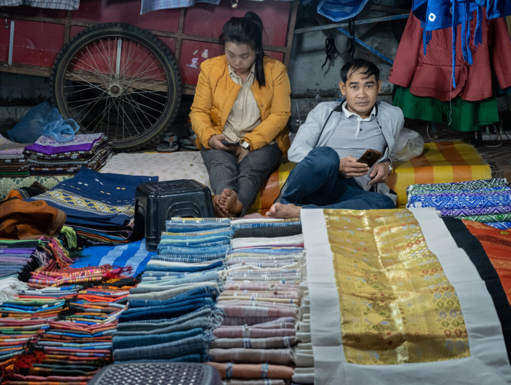 two people sitting behind a large number of fabric pieces for sale